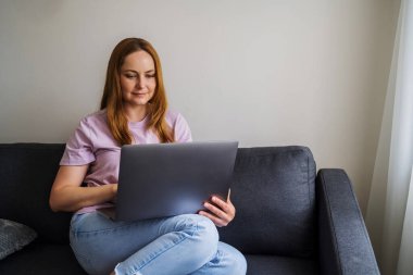 İnternette çalışan bir kadın, oturma odasında bir koltukta oturuyor ve dizüstü bilgisayar kullanıyor. 