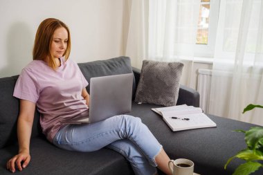 İnternette çalışan bir kadın, oturma odasında bir koltukta oturuyor ve dizüstü bilgisayar kullanıyor. 