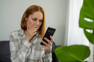 Kanepede oturan mutsuz kadın akıllı telefonundan kötü haberler okuyor.