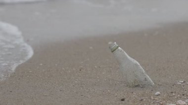 Kirlilik Konsepti: Cam şişe çöpü arka planda beyaz deniz dalgalarıyla kumsalda yatar.