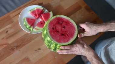 Adamın biri karpuzu ev mutfağında bıçakla parçalara ayırıyor. Ferahlatıcı bir yaz tatlısı manzarası.