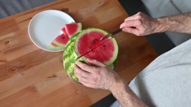 Adamın biri karpuzu ev mutfağında bıçakla parçalara ayırıyor. Ferahlatıcı bir yaz tatlısı manzarası.