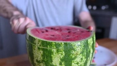 Adamın biri karpuzu ev mutfağında bıçakla parçalara ayırıyor. Ferahlatıcı bir yaz tatlısı manzarası.