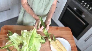 Kadın evdeki mutfakta yeşil yapraklardan taze mısır temizliyor. Ev yemeği, organik sarı mısır, sağlıklı sebze mevsimi. Yemek pişirmek için mısır koçanı temizleme işlemi