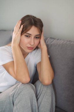 Kafasını tutan genç bir kadın, sinirli, modern oturma odasında kanepede oturuyor..