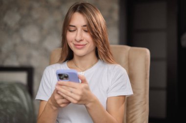 Genç bir kadın, yüzünde bir gülümsemeyle akıllı telefonuna odaklanmış rahat bir sandalyede oturmaktan zevk alır. Atmosfer rahat ve davetkar..