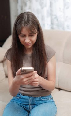 Genç bir kadın, yüzünde bir gülümsemeyle akıllı telefonuna odaklanmış rahat bir kanepede oturmaktan zevk alır. Atmosfer rahat ve davetkar..