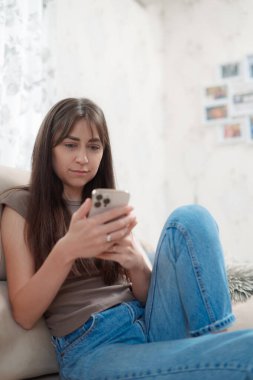 Genç bir kadın, yüzünde bir gülümsemeyle akıllı telefonuna odaklanmış rahat bir kanepede oturmaktan zevk alır. Atmosfer rahat ve davetkar..