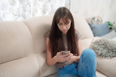 A young woman enjoys her time sitting in a cozy sofa, focused on her smartphone with a smile on her face. The atmosphere is relaxed and inviting. clipart