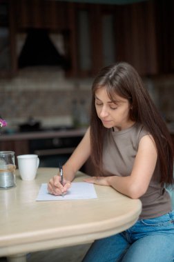 Genç bir kadın mutfakta bir masada oturuyor, kalemle bir kağıt parçasına dikkatlice yazıyor.