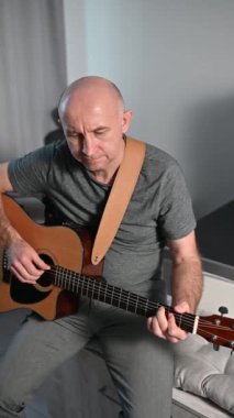 Modern koltukta otururken akustik gitar çalan müzisyen, minimalist yaşam alanındaki modern duvar lambalarıyla hafifçe aydınlatılıyor.