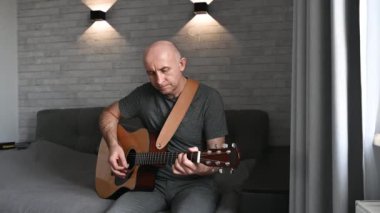 Modern koltukta otururken akustik gitar çalan müzisyen, minimalist yaşam alanındaki modern duvar lambalarıyla hafifçe aydınlatılıyor.