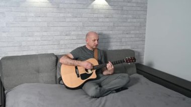 Modern koltukta otururken akustik gitar çalan müzisyen, minimalist yaşam alanındaki modern duvar lambalarıyla hafifçe aydınlatılıyor.