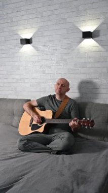 Modern koltukta otururken akustik gitar çalan müzisyen, minimalist yaşam alanındaki modern duvar lambalarıyla hafifçe aydınlatılıyor.