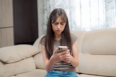 Genç bir kadın, yüzünde bir gülümsemeyle akıllı telefonuna odaklanmış rahat bir sandalyede oturmaktan zevk alır. Atmosfer rahat ve davetkar..