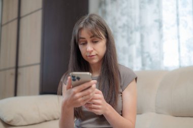 Genç bir kadın, yüzünde bir gülümsemeyle akıllı telefonuna odaklanmış rahat bir sandalyede oturmaktan zevk alır. Atmosfer rahat ve davetkar..