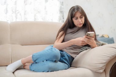 Genç bir kadın, yüzünde bir gülümsemeyle akıllı telefonuna odaklanmış rahat bir sandalyede oturmaktan zevk alır. Atmosfer rahat ve davetkar..