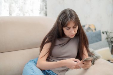 Genç bir kadın, yüzünde bir gülümsemeyle akıllı telefonuna odaklanmış rahat bir sandalyede oturmaktan zevk alır. Atmosfer rahat ve davetkar..