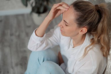 Genç bir kadın, gözleri kapalı bir kanepede oturuyor. Şakaklarını rahatsızlık içinde tutuyor. Çağdaş bir yaşam alanında baş ağrısı çektiğini gösteriyor..