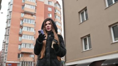Kürk mantolu, deri pantolonlu, botlu şık bir kadın. Şehir arka planında kahve içerken akıllı telefondan mesaj atıyor.
