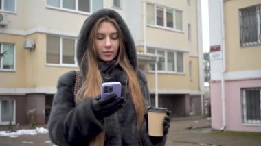 Kürk mantolu, deri pantolonlu, botlu şık bir kadın. Şehir arka planında kahve içerken akıllı telefondan mesaj atıyor.