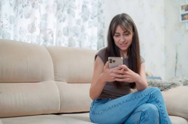 Genç bir kadın, yüzünde bir gülümsemeyle akıllı telefonuna odaklanmış rahat bir kanepede oturmaktan zevk alır. Atmosfer rahat ve davetkar..