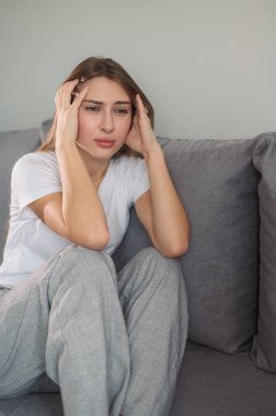 Kafasını tutan genç bir kadın, sinirli, modern oturma odasında kanepede oturuyor..