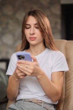 Genç bir kadın, yüzünde bir gülümsemeyle akıllı telefonuna odaklanmış rahat bir sandalyede oturmaktan zevk alır. Atmosfer rahat ve davetkar..