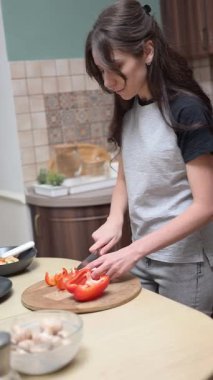 Genç bir kadın, ahşap bir kesme tahtasıyla kırmızı bir dolma biberi dilimliyor ve mutfağında yemek hazırlıyor.