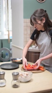 Genç bir kadın, ahşap bir kesme tahtasıyla kırmızı bir dolma biberi dilimliyor ve mutfağında yemek hazırlıyor.