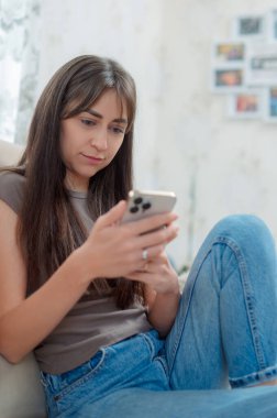 Genç bir kadın, yüzünde bir gülümsemeyle akıllı telefonuna odaklanmış rahat bir kanepede oturmaktan zevk alır. Atmosfer rahat ve davetkar..