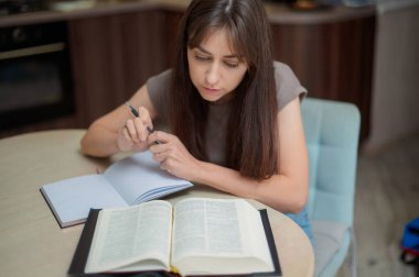 Bayan öğrenci çalışıyor ve evdeki sınava hazırlanıyor. Ders kitabını karıştıran ve notlar alan bir kadın.