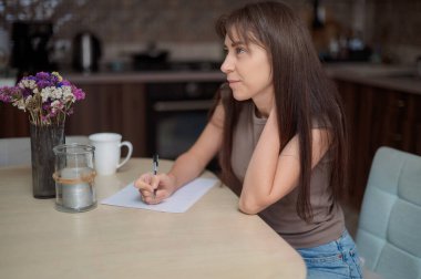 Genç bir kadın mutfakta bir masada oturuyor, kalemle bir kağıt parçasına dikkatlice yazıyor.