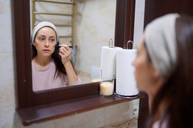 Bornoz ve saç bandı takan bir kadın banyo aynasının önünde fırçayla makyaj yapıyor.