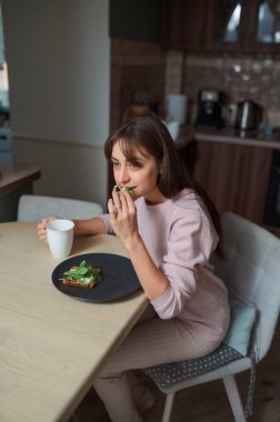 Genç bir kadın, modern mutfağında sebze ve çay eşliğinde sağlıklı bir tost kahvaltısı yapıyor.