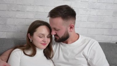 Genç çift, modern evlerindeki bir kanepede otururken sohbet ediyor ve el kol hareketi yapıyor. Birlikte vakit geçiriyorlar.