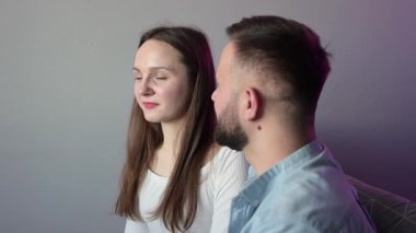 Genç çift, modern evlerindeki bir kanepede otururken sohbet ediyor ve el kol hareketi yapıyor. Birlikte vakit geçiriyorlar.
