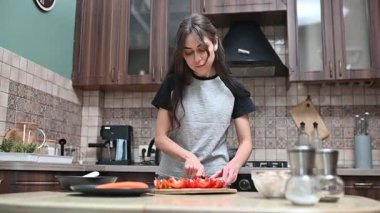 Genç bir kadın, ahşap bir kesme tahtasıyla kırmızı bir dolma biberi dilimliyor ve mutfağında yemek hazırlıyor.