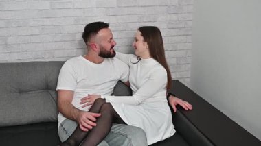Genç çift, modern evlerindeki bir kanepede otururken sohbet ediyor ve el kol hareketi yapıyor. Birlikte vakit geçiriyorlar.