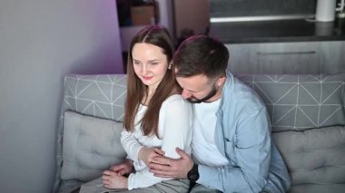 Romantik çift samimi bir anı paylaşıyor, kadın ve erkek modern dairelerinde bir kanepeye yaslanıyor, mor ortam aydınlatmasının yumuşak parıltısıyla banyo yapıyorlar.