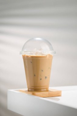 A glass of iced coffee with milk in a clear plastic cup on a white wooden table in a window-lit room. clipart