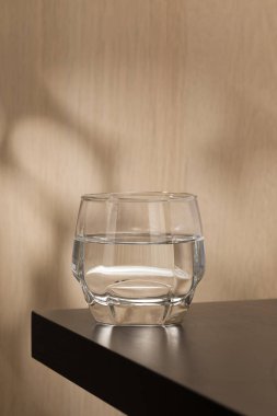 Temiz bir bardak içme suyu ahşap bir masada oturur, ışığı yansıtır ve ince bir gölge bırakır, sahnenin basitliğini ve saflığını vurgular..