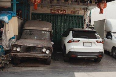 Bangkok \ Thailand - January 30 2025: A rusty truck and a new SUV parked side by side by a green gate, highlighting the juxtaposition of old and new. clipart