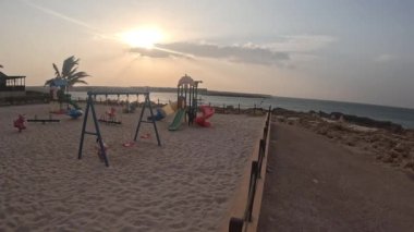 Gün batımında kumlu bir plajda salıncakta sallanan ve salıncaklar ve slaytlar da dahil olmak üzere oyun parkı ekipmanlarını gösteren bir çocuk, Umman 'daki Sur Sahili' nde akşam vakti rüzgarla hareket eden bir palmiye ağacıyla kumda konumlanıyor.