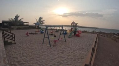 Sur, Umman 'da kumlu bir plajda, renkli oyun parkında salıncakla oynayan bir çocuk.
