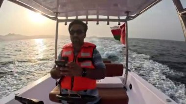 Güneş gözlüğü takan ve parlak turuncu can yeleği giyen bir adam, güvenli bir şekilde bir tekneyi sürüyor ve aynı zamanda cep telefonunu alıp Umman 'daki Muscat Wave' de tekne sürerken kullanıyor.