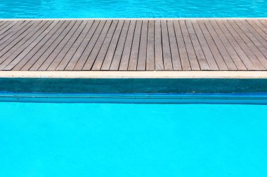 Arka planda ahşap döşemeli ve mavi suyu olan yüzme havuzu.