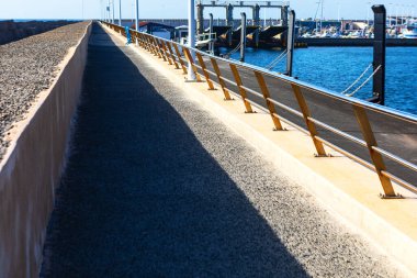 Korkuluğu ve arka planda rıhtımı olan uzun bir yürüyüş yolu. Geçit boş ve rıhtım teknelerle dolu. Uzağa uzanan beton yol
