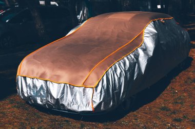 Arabanın üstü muşambayla kaplı. Araç muşambanın altına gizlenmiş, çimlerin üzerine park edilmiş.