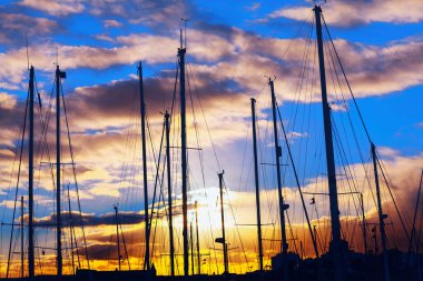 Alacakaranlık gibi limana demirlemiş yelkenliler. Gün batımında limanda yatların silueti. Bir grup yelkenli marinada sıralanır, güneş arka planda batar.